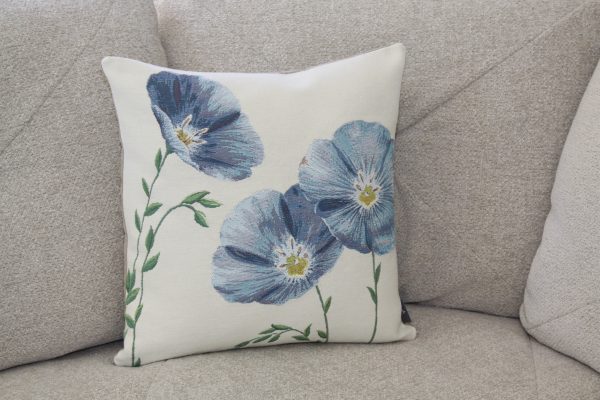 Coussin avec motif floral bleu, représentant des coquelicots, sur un canapé beige. Création artisanale en tissu jacquard.