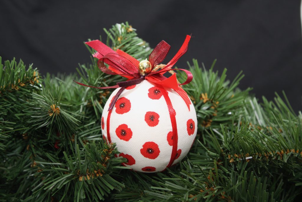 White fabric Christmas bauble adorned with red poppies, embellished with ribbons, suspended among fir branches.