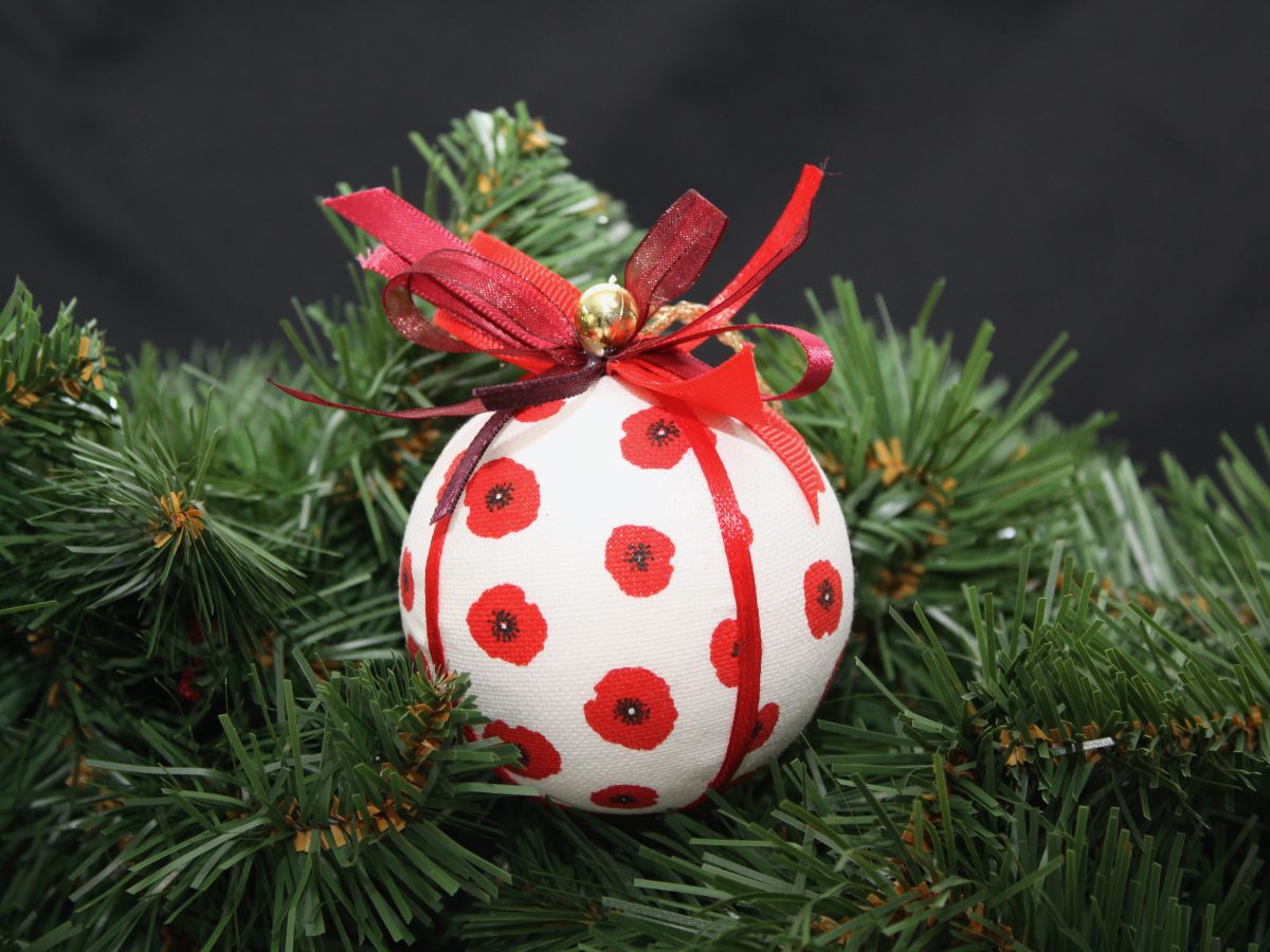 Boule de Noël en tissu blanc ornée de coquelicots rouges, agrémentée de rubans, suspendue parmi des branches de sapin.
