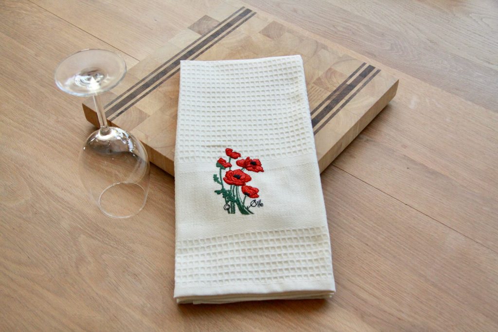 Torchon uni beige brodé de coquelicots, posé sur une planche en bois avec un verre à pied.