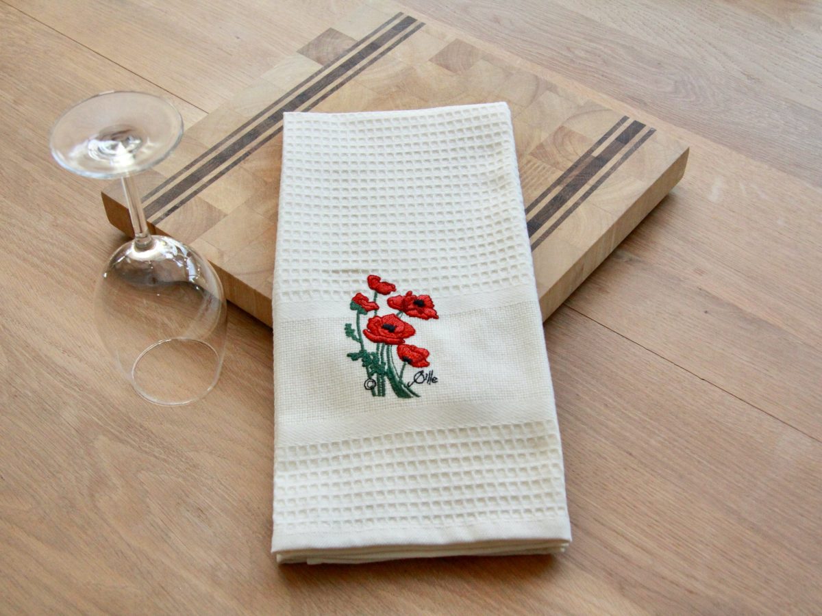 Torchon uni beige brodé de coquelicots, posé sur une planche en bois avec un verre à pied.