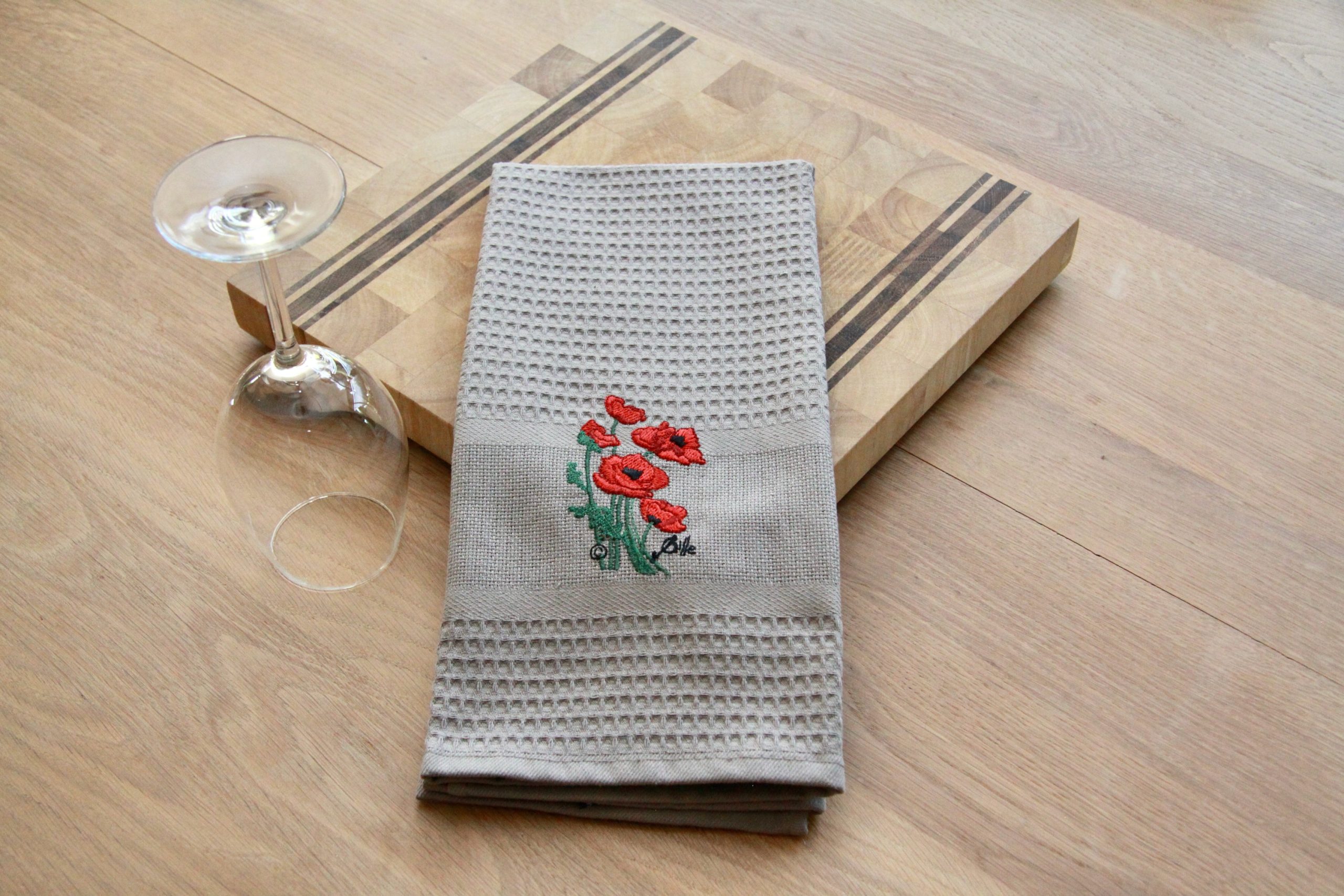 Torchon uni brodé avec des coquelicots, sur une planche en bois, avec un verre à pied à côté.