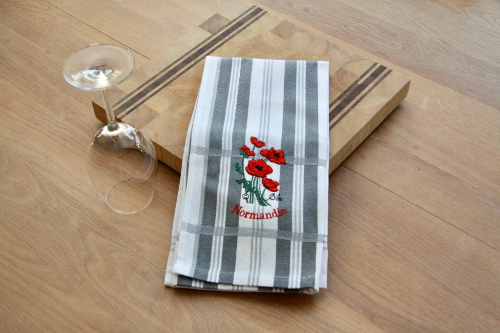 Torchon rayé brodé avec des coquelicots rouges, présenté sur une planche en bois, à côté d'un verre.