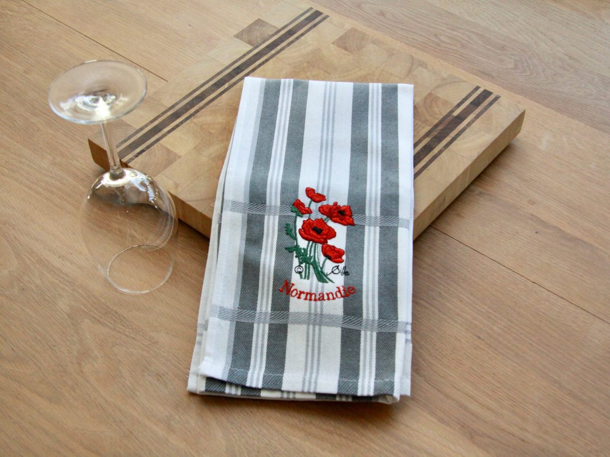 Torchon rayé brodé avec des coquelicots rouges, présenté sur une planche en bois, à côté d'un verre.